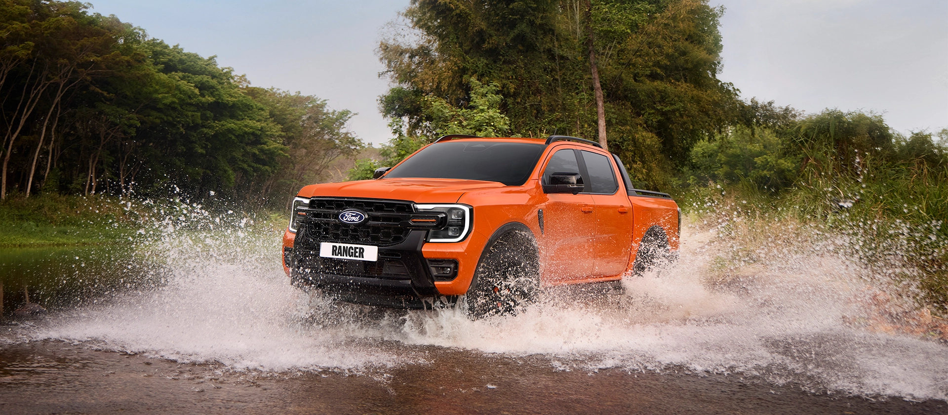Orange Ford Ranger driving through water on an off-road trail with splash effect.